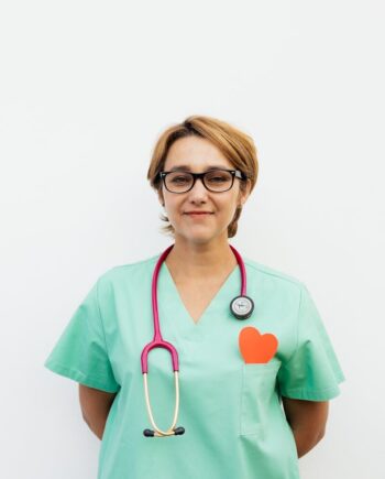 Portrait of a confident female doctor in green scrubs with a stethoscope and eyeglasses against a white background.