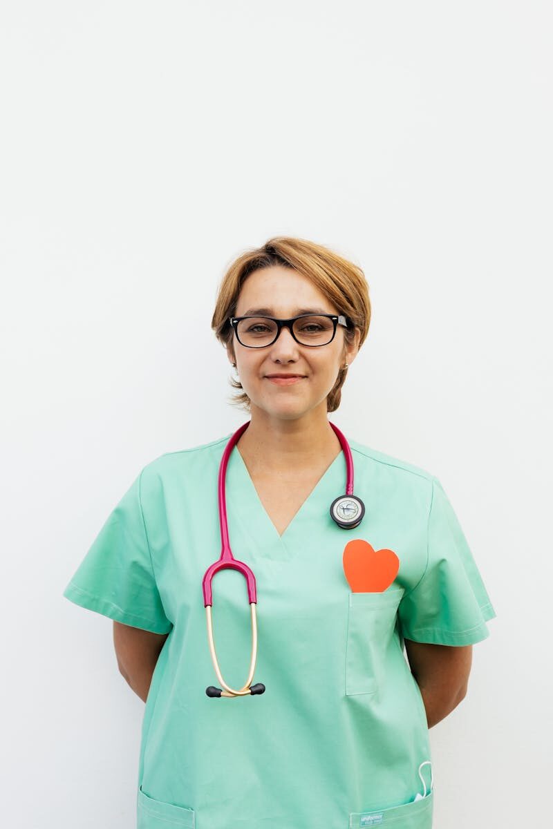 Portrait of a confident female doctor in green scrubs with a stethoscope and eyeglasses against a white background.
